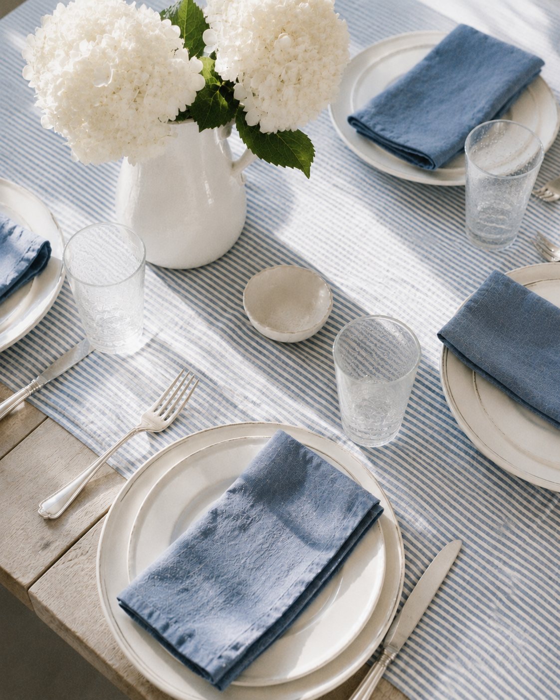 A sunlit table set with a blue-and-white striped runner, white plates, blue linen napkins and white hydrangea