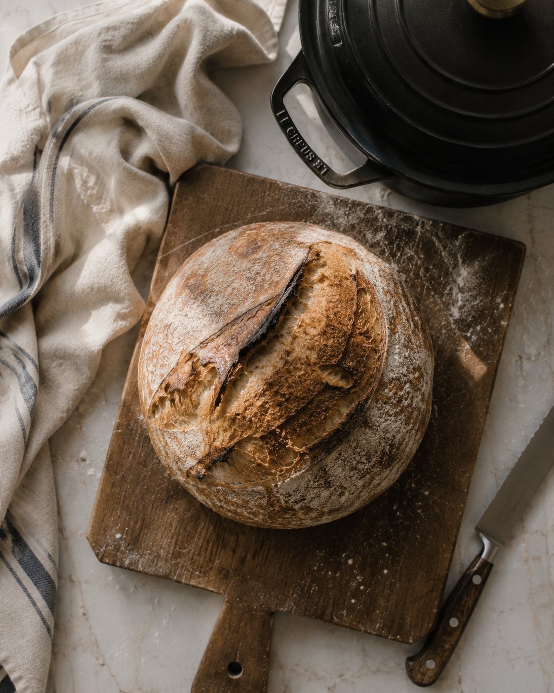 A rustic sourdough loaf on a floured wooden board beside a cast-iron Dutch oven