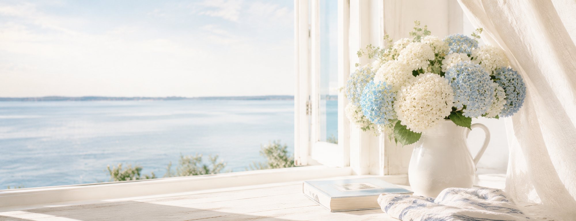 An open window with a sheer curtain looking out over calm blue water, white hydrangea on the sill